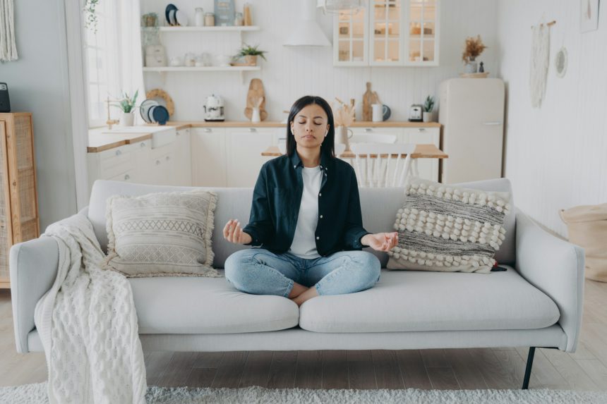 Female practice yoga, relax, sitting on sofa in lotus pose at home. Healthy lifestyle, stress relief