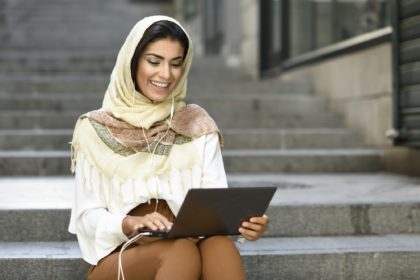 Spain, Granada, young muslim woman wearing hijab using laptop sitting on urban steps